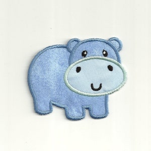 Può includere: Un'applique in feltro blu chiaro di un ippopotamo sorridente. L'ippopotamo ha un viso blu chiaro con occhi neri e un sorriso nero.