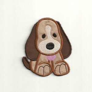 Könnte beinhalten: Gestickter Aufnäher in Braun und Beige, der einen Cartoon-Hund mit rosa Halsband zeigt. Der Hund sitzt mit hochgestreckten Pfoten.