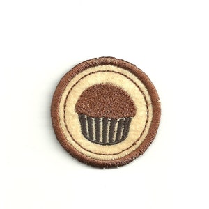 May include: A brown and white embroidered patch featuring a brown cupcake design. The cupcake has a brown top and a dark grey bottom with vertical lines.