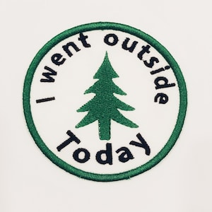 May include: A green and white embroidered patch with a green pine tree in the centre and the text "I went outside today" around the tree.