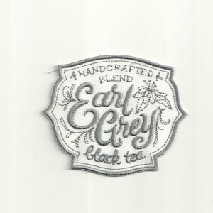 May include: A white fabric patch with gray embroidery. The patch features a stylized design of a flower and the text "A Handcrafted Blend Earl Grey Black Tea".