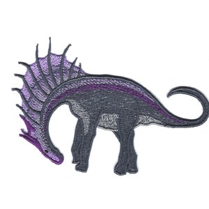 Può includere: Toppa ricamata di un dinosauro viola e grigio con una grande cresta spinosa. Il dinosauro è rivolto a sinistra e ha la coda arricciata.