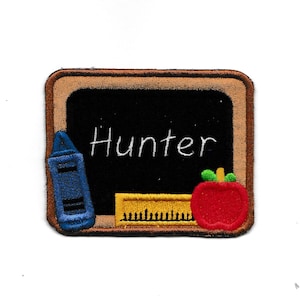 May include: A brown and black felt chalkboard with a white border. The chalkboard has a blue crayon, a yellow ruler, and a red apple on it. The name "Hunter" is written in white on the chalkboard.