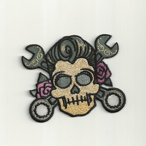 May include: Embroidered iron-on patch featuring a skull with a pompadour hairstyle, crossed wrenches, and pink roses. The skull is gold with black outlines, the wrenches are green with gold accents, and the roses are pink with green leaves.