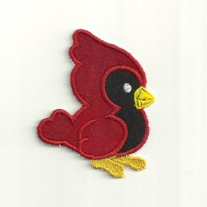 Puede incluir: Aplique de fieltro rojo en forma de pájaro cardenal con pico y patas amarillas. El pájaro está mirando hacia la derecha y tiene un ojo negro.