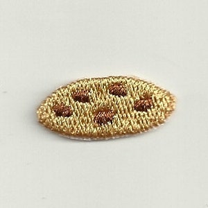May include: A small, embroidered patch in the shape of a cookie with brown chocolate chips on a beige background.