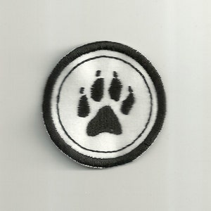 Könnte beinhalten: Runder, gestickter Aufnäher mit einem schwarzen Pfotenabdruck auf weißem Grund, umrandet von einem schwarzen Rand. Der Aufnäher ist zum Aufnähen oder Aufbügeln auf Textilien wie Kleidung oder Taschen gedacht.