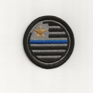 May include: A round black and gray embroidered patch with a thin blue line and a gold star in the center. The patch is surrounded by a black border.
