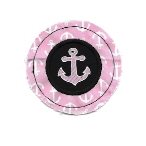 Pode incluir: Círculo de tecido rosa com um círculo preto no centro. O círculo preto tem uma âncora rosa no centro. O tecido rosa é coberto de âncoras brancas.