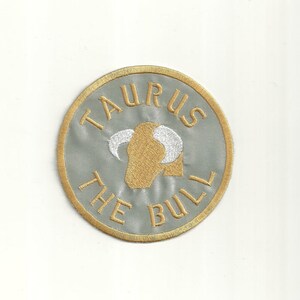 Könnte beinhalten: Ein gold gestickter Aufnäher mit grauem Hintergrund. Der Aufnäher zeigt einen Stierkopf mit weißen Hörnern und dem Text "Taurus The Bull".