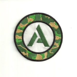 Op de afbeelding: Een geborduurde patch met een groen en bruin camouflagepatroon, met een witte cirkel met een groene letter "A" in het midden.