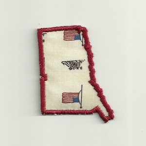 May include: A fabric patch in the shape of a state, with a red border. The patch features a cannon and two American flags. The background is off-white.