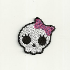 May include: White skull with black eyes and eyelashes, wearing a pink bow with purple polka dots.