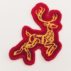 Puede incluir: Un parche de fieltro rojo con un diseño bordado en oro de un reno estilizado. El reno está corriendo y tiene grandes cuernos.