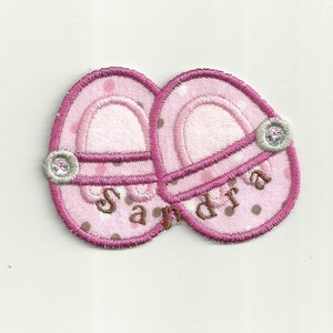 May include: Pink fabric applique of a pair of baby slippers with brown polka dots and the name "Sandra" embroidered on them.