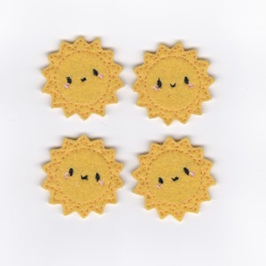 Puede incluir: Cuatro aplicaciones de fieltro amarillo en forma de sol con caras sonrientes bordadas. Cada sol tiene un rubor rosado en las mejillas y ojos negros.