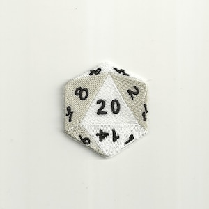 Op de afbeelding: Een witte en grijze geborduurde twintigzijdige dobbelsteen met zwarte cijfers. De dobbelsteen heeft de vorm van een dodecaëder.