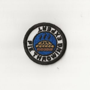 May include: Embroidered patch with a white background and a black border. The patch features a blue circle with a pie graphic and the words "PIE THROWING EXPERT". The pie is brown and gold, with steam rising from it.