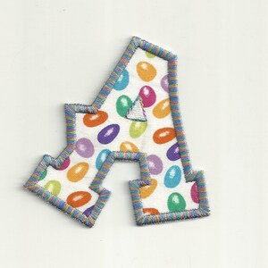 Könnte beinhalten: Gesticktes Stoff-Buchstaben-Patch "A" mit weißem Hintergrund, bedeckt mit bunten Gelee-Bohnen-Formen. Der Patch hat einen mehrfarbigen, genähten Rand. Ein kleines weißes Dreieck ist in der Mitte des Buchstabens aufgenäht.