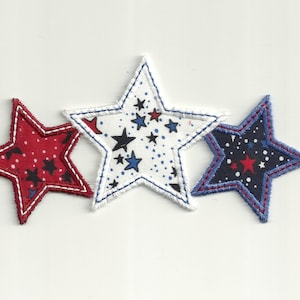 Op de afbeelding: Drie stoffen sterren met een witte achtergrond en rode, blauwe en zwarte sterren. De sterren zijn gestikt met wit, blauw en rood garen.