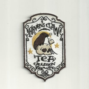May include: A brown and white embroidered patch with the text "Raven's Claw Tea Cauldron Brewed". The patch features a raven holding a cauldron and a skull with a spider web.