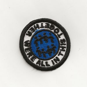 Könnte beinhalten: Ein gestickter Aufnäher in Schwarzweiß mit einem blauen Kreis in der Mitte. Der Kreis zeigt schwarze Strichmännchen, die sich an den Händen halten. Der Text um den Kreis herum lautet "We're All In This Together".