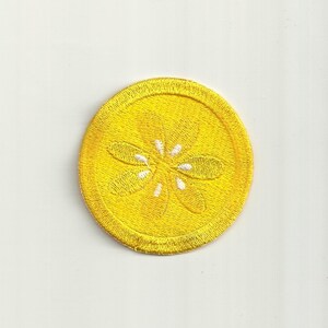 Puede incluir: Un parche amarillo bordado para planchar con un diseño de flor blanca. El parche es circular y tiene un borde amarillo.