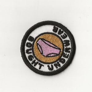 May include: A white and gold embroidered patch with black stitching. The patch is round and features a pink pair of underwear with the text "Bought Underwear" around the outside.