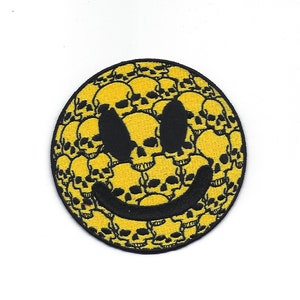 Puede incluir: Parche de cara sonriente amarillo con contorno negro. La cara sonriente está formada por un patrón de calaveras negras.
