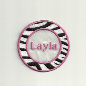 May include: A round, white and black zebra print iron-on patch with a pink border and the name "Layla" embroidered in pink inside a white circle.