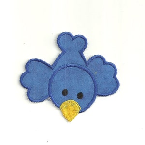 Puede incluir: Una aplicación de fieltro azul de un pájaro con un pico amarillo y ojos negros. El pájaro está mirando al espectador con las alas extendidas.