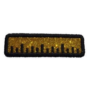 May include: A rectangular patch featuring a ruler design. The ruler's body is filled with gold glitter, and the edges are outlined in black. The ruler has black markings indicating measurements. The patch is likely for decorative purposes.