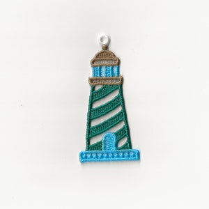 Puede incluir: Un adorno de faro bordado en verde y azul con un lazo blanco para colgar.