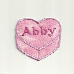 Puede incluir: Parche de fieltro rosa en forma de corazón con el nombre Abby bordado en hilo rosa.