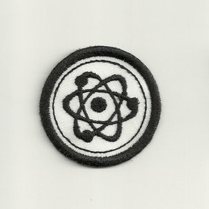 May include: A black and white embroidered patch with an atom symbol in the center. The atom symbol is surrounded by a white circle with a black border.