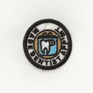 May include: A white embroidered patch with a black border. The patch features a tooth in a blue and gold container with the text "Made By A Dentist" and "Law Appt" around the border.