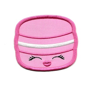 以下が含まれることがあります： ピンクのフェルトでできた、笑顔でウィンクしている漫画の顔のアップリケ。ピンクと白のストライプの帽子をかぶっています。目は黒く、口はピンクです。