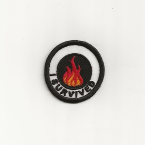 May include: A black and white embroidered patch with a red and orange flame in the center. The text "I Survived" is written around the flame in a circle.
