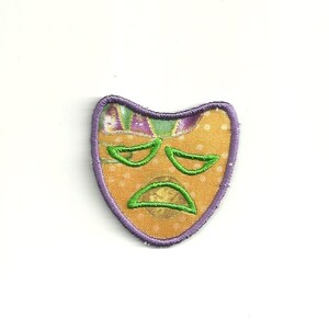 May include: Embroidered patch in the shape of a sad mask. The mask has a yellow background with white polka dots, green eyes and mouth, and a purple border. The top of the mask has a patterned design with purple and green colours.
