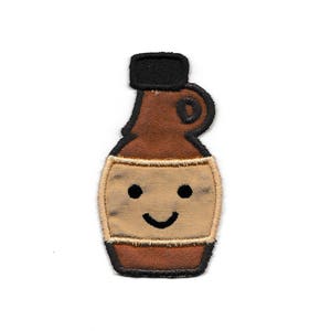 Pode incluir: Patch bordado com uma garrafa castanha com tampa preta e um rosto sorridente. O patch tem um rótulo bege e uma contorno preto. Adequado para roupas ou acessórios.