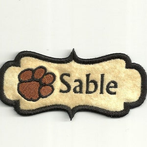 May include: A beige fabric patch with a black embroidered border. The patch features a brown paw print and the word "Sable" in black embroidery.