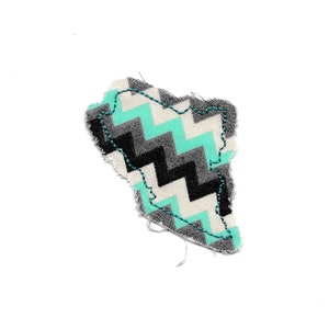 May include: A small, irregularly shaped fabric patch featuring a chevron design in black, white, and teal. The edges are finished with a teal zigzag stitch, set against a plain white background.