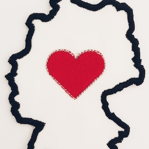 May include: A red heart embroidered on a white fabric background in the shape of the outline of Germany.