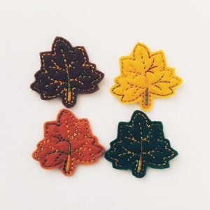 Puede incluir: Cuatro aplicaciones de fieltro en forma de hoja de arce en diferentes colores: marrón, amarillo, naranja y verde. Cada hoja tiene un diseño bordado detallado.