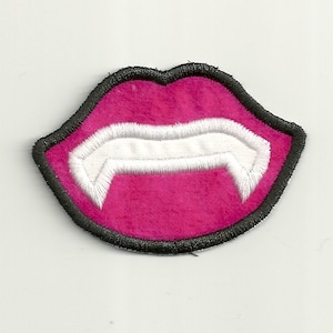 May include: A pink embroidered patch in the shape of a mouth with white fangs. The patch has a black border and is likely designed to be sewn or ironed onto clothing or accessories. The design is a playful take on a vampire theme.