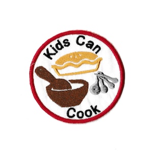 Peut inclure: Un écusson brodé circulaire blanc et rouge avec le texte "Kids Can Cook" en lettres noires. L'écusson représente un bol brun avec une cuillère en bois, une tarte jaune et trois cuillères en argent.