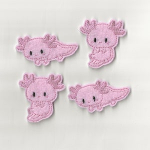 May include: Four pink felt axolotl patches with black embroidered eyes. The axolotls are in different poses, some with their limbs outstretched and others with their limbs tucked in.