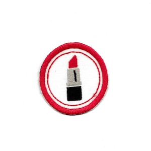 Pode incluir: Um remendo de ferro bordado com um design de batom. O batom é vermelho com uma faixa prateada e uma base preta.