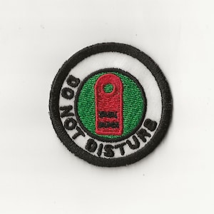 May include: A round, black and white embroidered patch with the text "Do Not Disturb" and a red door hanger with a white "Do Not Disturb" sign inside a green circle.