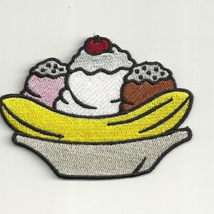 May include: An embroidered patch of a banana split ice cream sundae. The banana split is in a white bowl with a yellow banana on top. The ice cream is white, pink, and brown with chocolate chips. There is a red cherry on top of the white ice cream.